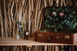Auf einem Holztisch steht ein offener Vintage-Koffer, gefüllt mit grünen Blättern und rosa Blumen, perfekt für eine Hochzeit. Eine kleine Vase mit Blumen, ein gerahmtes Bild und eine Kerze begleiten ihn vor einem Hintergrund aus vertikalen Holzzweigen.