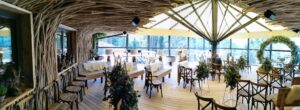 Ein geräumiger Innenbereich mit Holzboden und natürlichem, zweigähnlichem Dekor an Wänden und Decke, perfekt für Hochzeitsplanung. Er ist mit Holzstühlen und -tischen ausgestattet, die mit Blumenarrangements geschmückt sind. Große Fenster bieten einen Blick auf das Grün draußen.