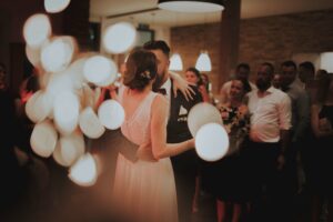 Braut und Bräutigam tanzen eng beieinander in einem warm beleuchteten Raum, umgeben von Gästen, die ihre Hochzeit feiern. Weiche, unscharfe Lichter im Vordergrund schaffen eine romantische Atmosphäre. Die Braut trägt ein weißes Kleid und der Bräutigam einen dunklen Anzug, was elegante Hochzeitsplanung verkörpert.
