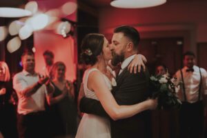 Braut und Bräutigam küssen sich während ihres Hochzeitsempfangs auf der Tanzfläche. Die Braut, die einen Blumenstrauß hält, ist von enthusiastisch klatschenden Gästen umgeben. Der Raum strahlt mit seinem roten und sanften Ambientelicht Wärme aus und spiegelt ihre wunderschön geplante Hochzeitsplanung perfekt wider.