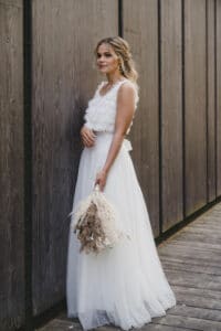Eine Frau in einem weißen ärmellosen Kleid, die die Eleganz einer Hochzeit verkörpert, steht an einer Holzwand und hält einen Strauß getrockneter Blumen in der Hand. Mit gestyltem blondem Haar blickt sie leicht in die Ferne. Die Außenkulisse auf dem Holzdeck trägt zur ruhigen Atmosphäre bei, perfekt für Inspiration bei der Hochzeitsplanung.