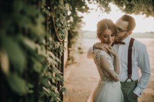 Ein Paar umarmt sich auf einem sonnenbeschienenen Weg und feiert seine Hochzeit. Der Bräutigam, in Hosenträgern und Fliege, küsst die Schläfe der Braut, während sie in ihrem langärmeligen Spitzenbrautkleid sanft lächelt. Umgeben von Grün an diesem warmen, ruhigen Tag ist ihre Liebe so perfekt, wie es sich jede Hochzeitsplanung nur wünschen kann.