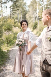 Ein Paar schlendert Hand in Hand einen sonnigen Waldweg entlang und träumt von seiner Hochzeit. Die Frau, in einem traditionellen Kleid mit einem Blumenstrauß, lächelt freudig. Der Mann, in Weste und Hemd gekleidet, blickt sie herzlich an. Üppiges Grün umgibt sie an diesem perfekten Tag zum Heiraten.