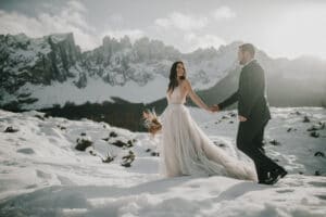 Braut und Bräutigam gehen Hand in Hand durch die schneebedeckten Berge, ihre Hochzeit wird von dramatischen Felsgipfeln eingerahmt. Die Braut in einem fließenden weißen Kleid mit getrockneten Blumen in der Hand strahlt, als das Sonnenlicht durch die Wolken fällt.