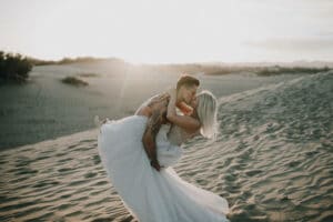 Ein Paar umarmt sich leidenschaftlich in einer Wüste bei Sonnenuntergang und fängt einen magischen Hochzeitsmoment ein. Der Mann hebt die Frau hoch, deren fließendes weißes Kleid im Wind weht. Sanddünen erstrecken sich unter einem klaren Himmel in die Ferne und bilden die perfekte Kulisse für ihre Liebesgeschichte.