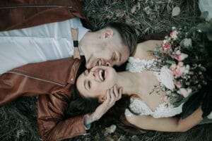 Ein glückliches Paar liegt im Gras, der Mann trägt eine braune Jacke und Fliege, die Frau ein Spitzenkleid und hält einen Strauß rosa und weißer Blumen in der Hand. Sie lächeln und umarmen sich zwischen trockenen Blättern und genießen ihr Hochzeitsglück.