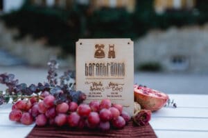 Eine Getränkekarte aus Holz mit Abbildungen eines Telefons und eines Staubsaugers listet Bargetränke auf, perfekt für Ihre Hochzeitsplanung. Sie liegt auf einem Tisch, der mit roten Trauben, geschnittenen Granatäpfeln und Eukalyptusblättern geschmückt ist, während der Hintergrund eine romantische Unschärfe ausstrahlt.