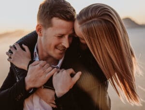 Ein Paar umarmt sich im Freien, wobei die Frau den Mann von hinten umarmt und ihr Kinn auf seine Schulter legt. Sie wirken glücklich und träumen von ihrer zukünftigen Hochzeit. Sie tragen legere Jacken. Im Hintergrund geht die Sonne unter und wirft ein warmes Licht.