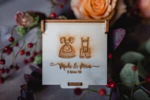 Eine Holzkarte mit eingravierten Bildern von Trachten und dem Text „Michi & Manu, 21. Oktober 2018“. Umgeben von Deko-Elementen für eine bezaubernde Hochzeit, darunter Rosen, Beeren und Eukalyptusblätter.