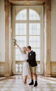 Ein Paar küsst sich vor einem großen Bogenfenster und drückt damit die Freude ihres Hochzeitstages aus. Die Frau hält einen Blumenstrauß hoch, beide sind halbformell gekleidet, inmitten kunstvoller Dekoration und eines karierten Bodens, perfekt für ihre elegante Hochzeitsfeier.
