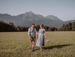 Ein Paar in traditioneller bayerischer Kleidung geht Hand in Hand über eine Wiese mit üppigen grünen Bäumen und Bergen im Hintergrund und genießt die Freude des Heiratens. Die Frau hält einen Blumenstrauß und beide schauen sich an ihrem besonderen Tag strahlend an.