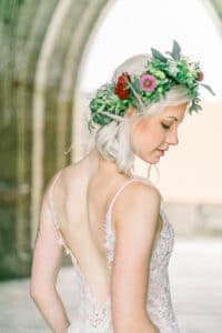 Eine Braut mit Blumenkrone und Spitzenkleid posiert anmutig in einem historischen Steinbogen. Ihr blondes Haar ist locker gestylt und sie blickt nach unten, was eine heitere und elegante Atmosphäre schafft und die perfekte Vision einer Traumhochzeit verkörpert.