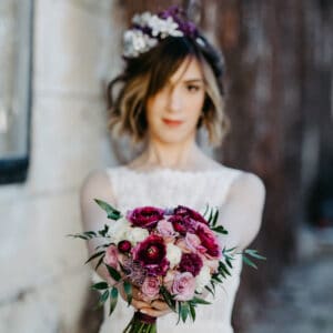 Eine Braut in einem ärmellosen weißen Kleid hält einen Blumenstrauß mit rosa, violetten und roten Blüten in der Hand und fängt damit die Essenz einer Hochzeit ein. Sie steht vor einem unscharfen Hintergrund, was dem Bild eine künstlerische und romantische Note verleiht, die perfekt zu jeder Hochzeitsplanung passt.