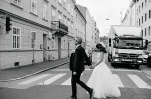 Braut und Bräutigam gehen Hand in Hand über eine Stadtstraße und verkörpern damit die Freude ihrer Hochzeit. Die Braut trägt ein fließendes weißes Kleid und der Bräutigam einen dunklen Anzug. Sie passieren Gebäude und einen weißen Lastwagen auf einem Zebrastreifen, alles wunderschön in Schwarzweiß festgehalten.