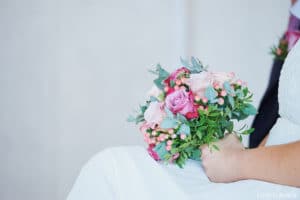 Eine Person in einem weißen Kleid hält einen Strauß rosafarbener und weißer Blumen gemischt mit Grün und verkörpert damit die Essenz einer Hochzeit. Der Hintergrund ist sanft verschwommen und hebt den Strauß hervor.