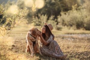 Eine Frau in einem geblümten Kleid und Hut, die wahrscheinlich gerade ihre Hochzeit plant, sitzt im Gras neben einem Hund und berührt zärtlich sein Gesicht. Sie genießen den sonnigen, üppigen Park mit einem kleinen Teich in der Nähe, umgeben von Bäumen – ein ruhiger Moment, perfekt für Inspiration bei der Hochzeitsplanung.