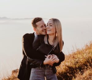 Ein Paar steht umarmt auf einem grasbewachsenen Hügel und denkt an die bevorstehende Hochzeit. Der Mann, der eine schwarze Jacke trägt, umarmt die Frau von hinten, während sie in ihrem schwarzen Oberteil und Jeans breit lächelt. Die dunstige Landschaft und das sanfte Licht schaffen eine heitere Kulisse für ihren gemeinsamen Traum vom Heiraten.