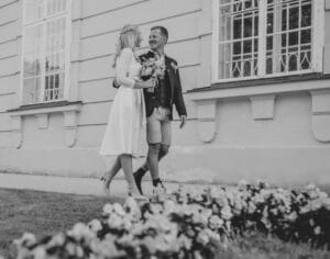 Ein Paar in formeller Kleidung lächelt sich an, während es auf einem Weg geht und verkörpert die Freude der Hochzeit. Die Frau hält einen Blumenstrauß in der Hand, was ihrem Moment Charme verleiht. Sie stehen vor einem Gebäude mit großen Fenstern und im Vordergrund ist eine Reihe Blumen zu sehen. Das Bild ist schwarzweiß.