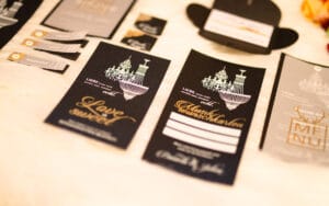 Eine Sammlung eleganter Hochzeitspapeterie für Ihre Hochzeit auf weißem Untergrund, mit schwarzen Einladungen mit goldenem und weißem Text und Kronleuchterillustrationen. Die Artikel umfassen ein Menü, Namensschilder und RSVP-Karten, die alle darauf ausgelegt sind, Ihrer Hochzeitsfeier einen Hauch von Luxus zu verleihen.
