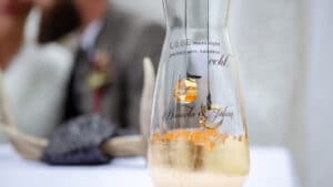 Eine Glasvase mit goldenen Ringen im Inneren und der deutschen Aufschrift „Liebe muss nicht perfekt sein, sondern echt“ verleiht der Hochzeit einen Hauch von Eleganz. Im Hintergrund sitzt eine verschwommene Figur an einem mit Geweihen geschmückten Tisch und vermittelt den Eindruck einer intimen Hochzeitsplanung.
