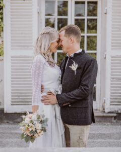 Vor einem Gebäude mit weißen Türen umarmt sich ein Paar. Die Frau, die ein weißes Kleid trägt und einen Blumenstrauß hält, steht mit ihrem Partner bei den Vorbereitungen für ihre Hochzeit. Er trägt eine dunkle Jacke und helle Hosen, während sie sich herzlich anlächeln und in ihre Hochzeitsplanung vertieft sind.