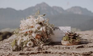 Ein Strauß weißer und rötlicher Blumen mit Grünpflanzen liegt auf einer Steinoberfläche neben einer Karte und einem kleinen Sukkulentenarrangement. Im Hintergrund erheben sich Berge unter einem klaren Himmel und bilden die perfekte Kulisse für die Hochzeitsplanung.