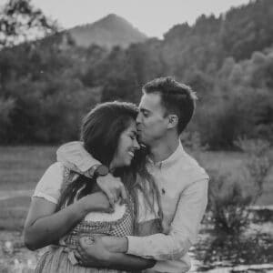 Ein Schwarzweißfoto eines Paares, das sich im Freien umarmt und einen zärtlichen Moment inmitten ihrer Hochzeitsplanung festhält. Der Mann küsst die Stirn der Frau, während beide lächeln. Sie stehen auf einem Feld mit Bergen und Bäumen im Hintergrund.