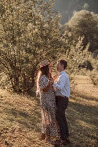 Auf einer sonnenbeschienenen Wiese, umgeben von Bäumen, lächelt sich ein Paar an, eingehüllt in die warme Glut ihres Hochzeitstages. Die Frau trägt ein langes geblümtes Kleid und einen Hut, während der Mann ein weißes Hemd und dunkle Hosen trägt, was eine mühelos romantische Atmosphäre schafft.