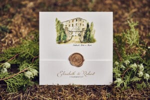 Eine Hochzeitseinladung zeigt die Abbildung eines umzäunten Gebäudes, umgeben von Bäumen. Versiegelt mit einem Wachssiegel mit den Initialen „E“ und „R“ steht darauf „Elisabeth & Robert, 8. September 2021“, elegant platziert auf einer natürlichen, moosbewachsenen Oberfläche.