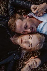 Ein Paar liegt auf trockenem Gras und träumt von seinem Hochzeitstag. Der Mann legt seinen Kopf auf die Schulter der Frau. Beide tragen schwarze Lederjacken, halten Händchen und stellen sich eine Zukunft vor, in der ihre enge und entspannte Verbindung zu Hochzeiten erblüht.