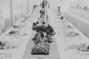 Ein langer, elegant gedeckter Esstisch ist mit einem Blumenschmuck und sorgfältig arrangierten Gläsern und Servietten geschmückt – perfekt für eine Hochzeit. Das Schwarz-Weiß-Bild fängt eine einladende und anspruchsvolle Atmosphäre ein und spiegelt eine sorgfältige Hochzeitsplanung wider.
