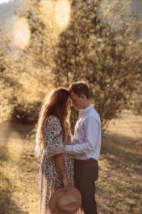 Auf einem sonnenbeschienenen Feld, wo Bäume als stumme Zeugen stehen, steht ein Paar eng beieinander und berührt sanft die Stirn. Die Frau hält einen Hut in einer Hand und verkörpert damit die Ruhe ihres Hochzeitstages, während sanftes Sonnenlicht hindurchfällt und ihren Moment der Liebe in einen magischen Glanz taucht.