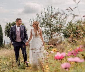 Braut und Bräutigam gehen Händchen haltend durch eine mit Wildblumen übersäte Wiese und verkörpern damit die Essenz einer perfekten Hochzeit. Die Braut trägt ein weißes Kleid, während der Bräutigam einen Anzug mit Weste trägt. Bäume und ein bewölkter Himmel bilden die malerische Kulisse für diesen idyllischen Moment ihrer Hochzeitsplanung.