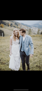 Braut und Bräutigam gehen Arm in Arm auf einem grasbewachsenen Hügel und feiern ihre Hochzeit. Die Braut trägt ein weißes Kleid, während der Bräutigam einen hellblauen Anzug trägt. Sanfte Hügel und Bäume erstrecken sich unter einem bewölkten Himmel in die Ferne und tragen zur ruhigen Kulisse ihres besonderen Tages bei.