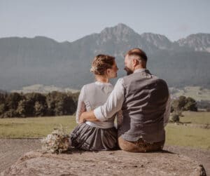 Ein Paar sitzt auf einem großen Felsen, umarmt sich und blickt sich inmitten einer Berglandschaft an. Die Frau hält einen Blumenstrauß in der Hand. Ihre traditionelle Kleidung passt perfekt zu ihrer Hochzeit, vor dem Hintergrund üppiger grüner Felder und entfernter Hügel.