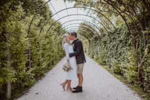 Ein Paar küsst sich unter einem gewölbten Spalier in einem üppigen, grünen Garten. Die Frau in einem weißen Kleid hält als Teil ihrer Hochzeit einen Blumenstrauß in der Hand, während der Mann einen Blazer und Shorts trägt. Sie sind von dichtem Laub auf einem Sandweg umgeben.