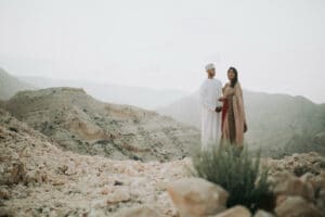 Inmitten einer felsigen Wüstenlandschaft steht ein Paar in traditioneller Kleidung und verkörpert den Geist des Heiratens. Der Mann trägt einen weißen Thobe und eine Kumma, während die Frau sich in einen gemusterten Schal hüllt. In der Ferne erstrecken sich trockene Berge unter einem blassen Himmel und bieten eine unvergessliche Kulisse für diesen besonderen Moment.