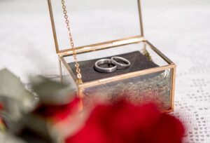 Ein Paar Silberringe in einer kleinen, offenen Glasbox mit Goldrahmen, platziert auf einer Oberfläche aus weißer Spitze – eine perfekte Note für jede Hochzeit. Der Vordergrund ist verschwommen und zeigt unscharfe rote Blumen.