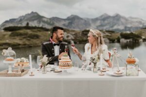 Ein Paar in Hochzeitskleidung sitzt an einem wunderschön dekorierten Tisch und verkörpert die Kunst der Hochzeitsplanung. Sie stoßen mit Weingläsern vor einer atemberaubenden Kulisse aus Bergen und einem See an, umgeben von einer Reihe von Desserts und Blumen – ein perfekter Moment, den sie bei der Feier ihrer Hochzeit in Erinnerung behalten sollten.