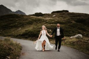 Eine Braut und ein Bräutigam laufen fröhlich Hand in Hand eine kurvenreiche Straße in einer Berglandschaft entlang und verkörpern damit den Geist einer Hochzeit. Das fließende weiße Kleid und der Brautstrauß bilden einen schönen Kontrast zum schwarzen Smoking des Bräutigams vor einem dramatischen, wolkigen Himmel.