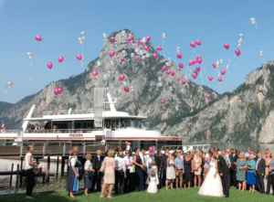 Eine Hochzeit am Seeufer mit Bergkulisse bietet die perfekte Kulisse. Die Gäste lassen rosa und weiße Luftballons in den Himmel steigen, während die „Poseidon“ in der Nähe ankert. Braut und Bräutigam, die die Freude verkörpern, stehen umgeben von ihren Lieben und feiern diesen besonderen Moment der Hochzeit.