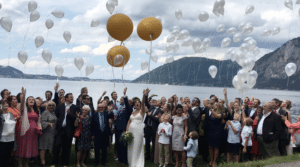 Eine Hochzeitsfeier mit einer großen Gruppe, die weiße und goldene Luftballons am Seeufer steigen lässt. Gäste jeden Alters sind in formelle Kleidung gekleidet und Berge und ein bewölkter Himmel dienen als Kulisse. Braut und Bräutigam stehen in der Mitte und genießen ihren unvergesslichen Hochzeitsmoment.