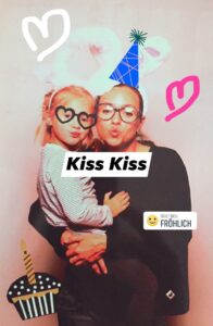 Eine Frau und ein Kind posieren verspielt, mit Hasenohren und einem Partyhut-Motiv. Das Kind trägt eine herzförmige Brille. Zu den Kritzeleien gehören Herzen, ein Kuchen und der Text „Kiss Kiss“, „Fühlt sich: Fröhlich“ mit einem lächelnden Emoji – ein Ausdruck der Freude, die die Hochzeitsplanung mit sich bringt.