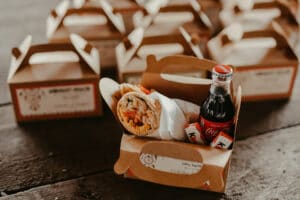 Eine Essensbox aus Pappe mit einem Wrap-Sandwich, einer kleinen Flasche Coca-Cola und einer Packung Ketchup steht auf einem Holztisch – perfekt für zwanglose Zusammenkünfte oder auch entspannte Hochzeitsplanungssitzungen. Im Hintergrund sind weitere ähnliche Boxen zu sehen.