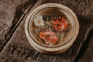 Eine runde Holzbox mit transparentem Deckel zeigt im Inneren zwei ineinander verschlungene Ringe auf rosa Blütenblättern und Trockenblumen. Der Text lautet „WE DO“ mit „Valentina & Frederic 8. Mai 2021“ darunter, perfekt für Ihre Hochzeit. Die Box ruht elegant auf einer rustikalen Holzoberfläche.