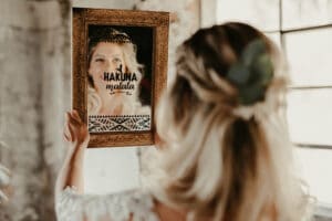 Eine Frau mit blondem Haar und einem grünen Accessoire hält einen rechteckigen, kunstvoll gerahmten Spiegel hoch. In der Spiegelung steht in dekorativen Lettern „Hakuna Matata“, was ihr Gesicht teilweise verdeckt – die perfekte Stimmung für eine stressfreie Hochzeitsplanung. Der Hintergrund ist sanft verschwommen.
