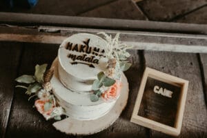 Eine rustikale zweistöckige weiße Torte mit „Hakuna Matata“-Dekoration, geschmückt mit Grünpflanzen und pfirsichfarbenen Rosen, steht auf einem Holzsockel – perfekt für eine Hochzeit. Daneben steht auf einem dunklen Holztisch ein gerahmtes Schild mit der Aufschrift „Torte“.