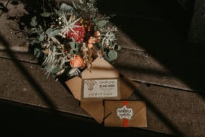Eine rustikale Hochzeitseinladungsserie liegt auf einem Holzboden, begleitet von einem Strauß getrockneter Blumen. Auf der Einladung steht in eleganter Schrift „Save the Date, 8. Mai 2021“. Schatten und Licht schaffen eine warme, natürliche Atmosphäre, perfekt für eine unvergessliche Hochzeitsfeier.