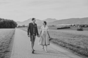 Ein Paar in traditioneller Kleidung hält sich an den Händen und geht eine Landstraße entlang, umgeben von weiten Feldern und fernen Bergen unter einem klaren Himmel. Die Szene ist friedlich und fängt eine zeitlose Verbindung ein, die sich wie der Beginn ihrer gemeinsamen Hochzeitsreise anfühlt.