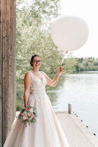 Eine Braut im weißen Brautkleid steht neben einer Holzmauer in der Nähe eines Sees und hält einen großen weißen Ballon und einen Strauß rosa und weißer Blumen in der Hand. Lächelnd vor der perfekten Hochzeitskulisse spiegelt die Szene mit Bäumen und Wasser als ruhigem Hintergrund eine sorgfältige Hochzeitsplanung wider.